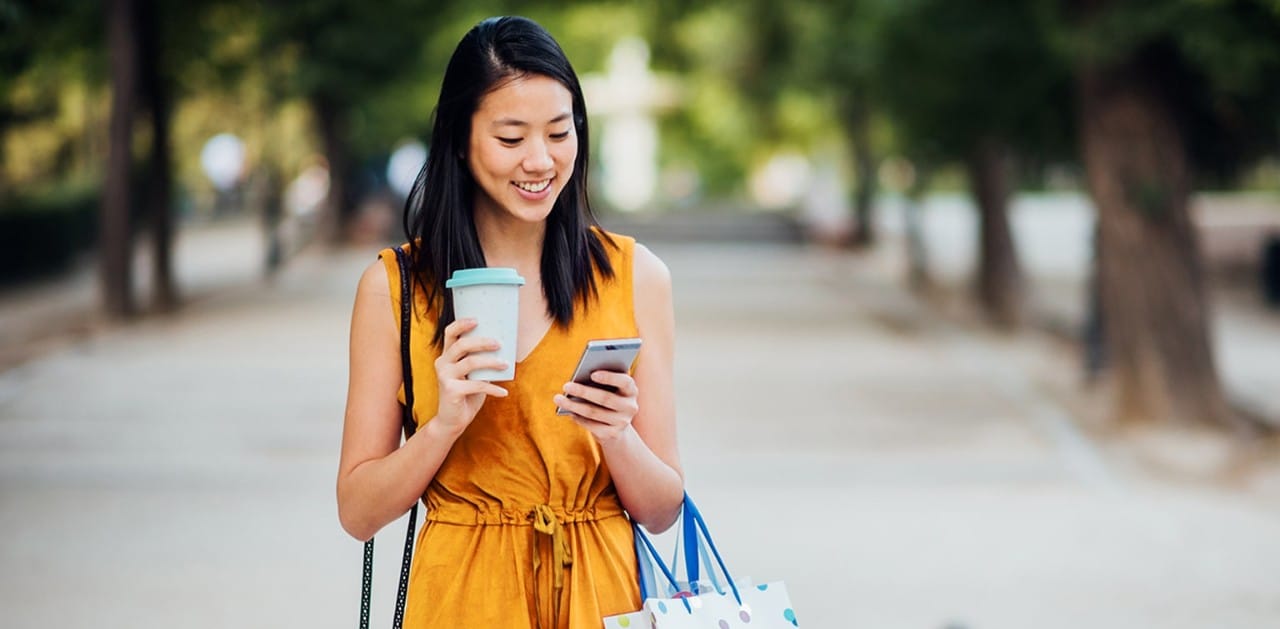Young Asian woman holding coffee cup and shopping bags and looking at smartphone