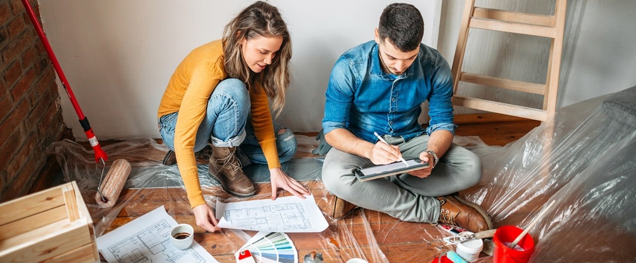 Young couple sitting on floor during home renovation looking at blueprint and table