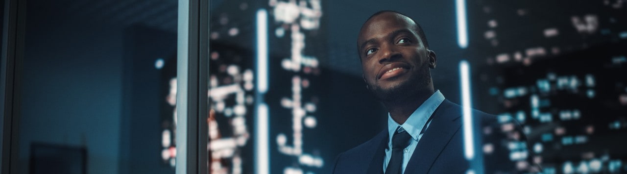 Happy, young African-American businessman looking out window at city at night