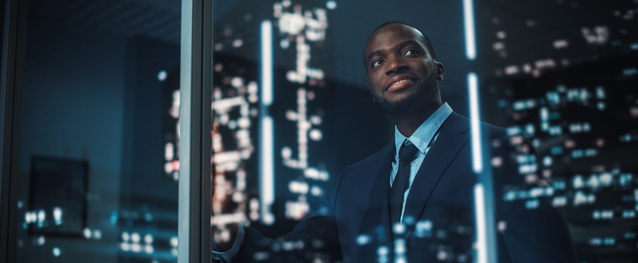 Happy, young African-American businessman looking out window at city at night
