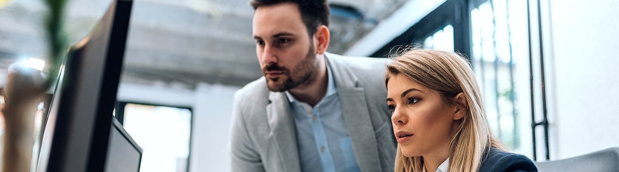 Business woman sitting with male co-worker standing next to her, both looking at a computer monitor