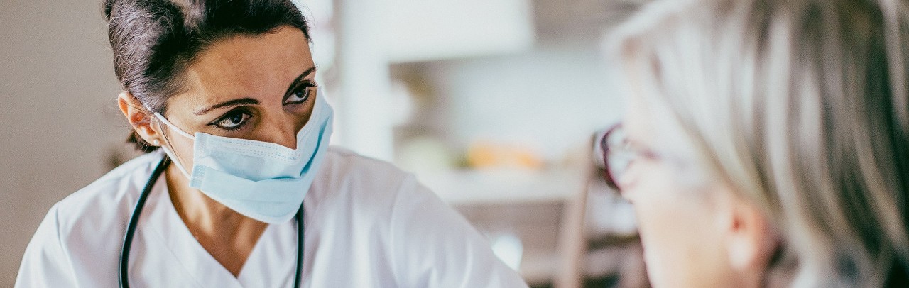 A medical professional in scrubs in mask with a stethoscope around her neck observing a patient.