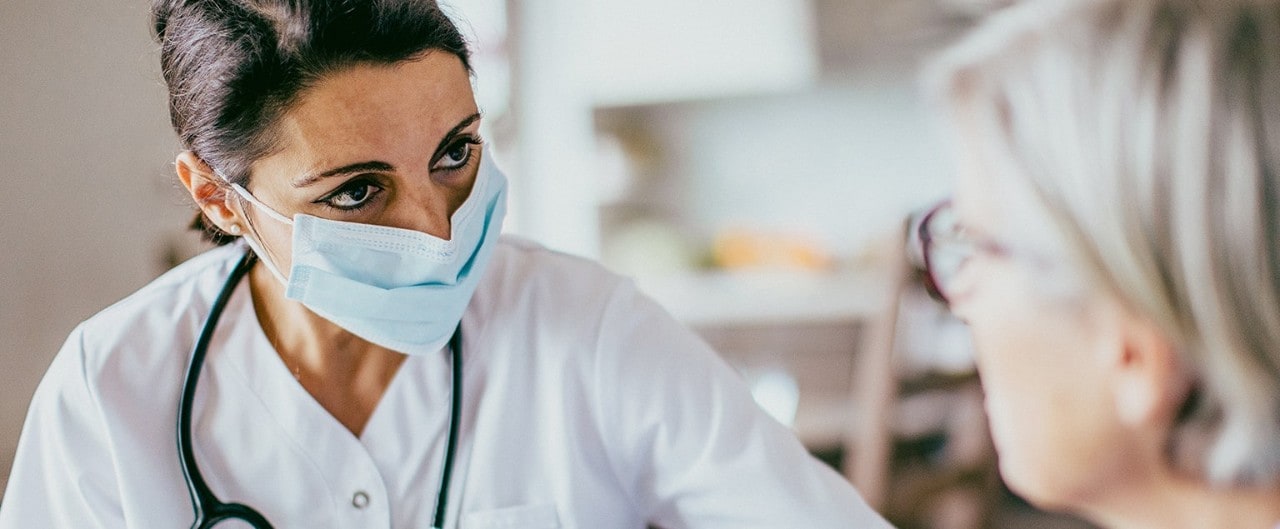 A medical professional in scrubs in mask with a stethoscope around her neck observing a patient.