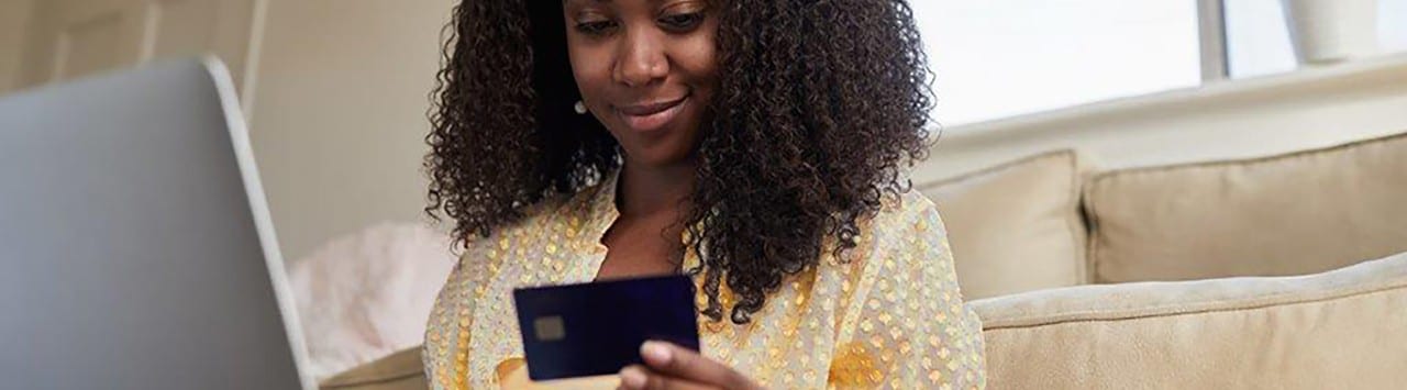 African-American woman leaning against couch holding credit card and laptop