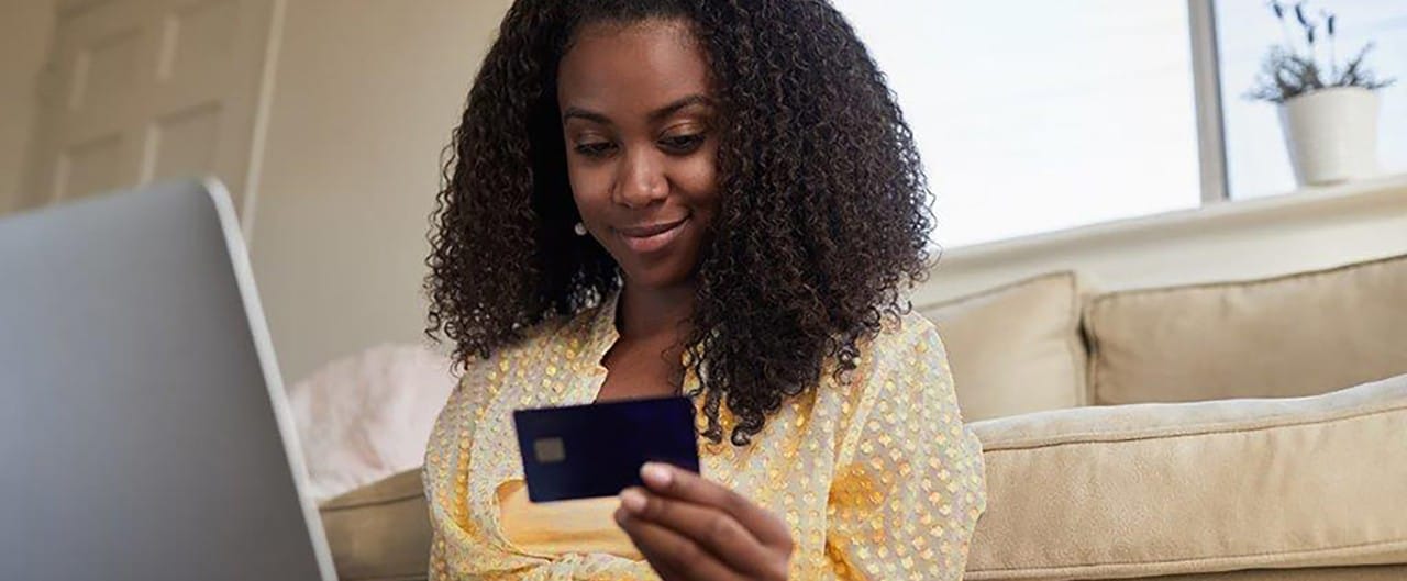 African-American woman leaning against couch holding credit card and laptop