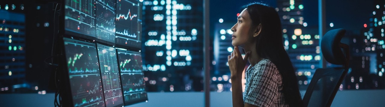 Asian female financial analyst reviewing stock data on multiple monitors