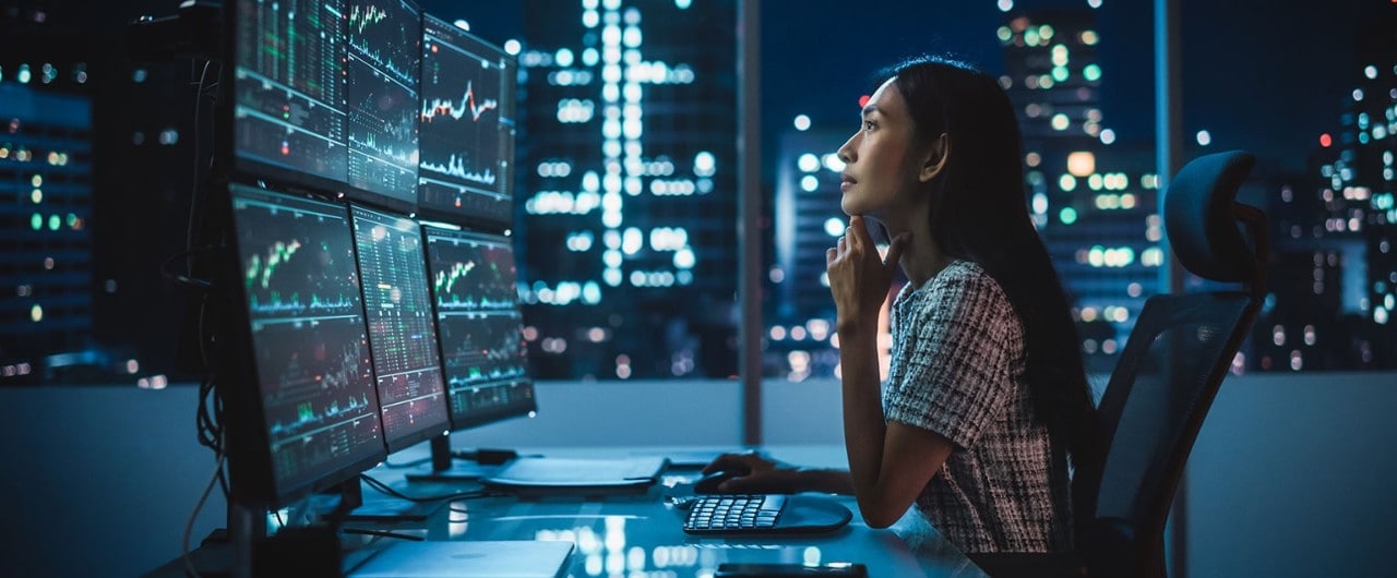 Asian female financial analyst reviewing stock data on multiple monitors