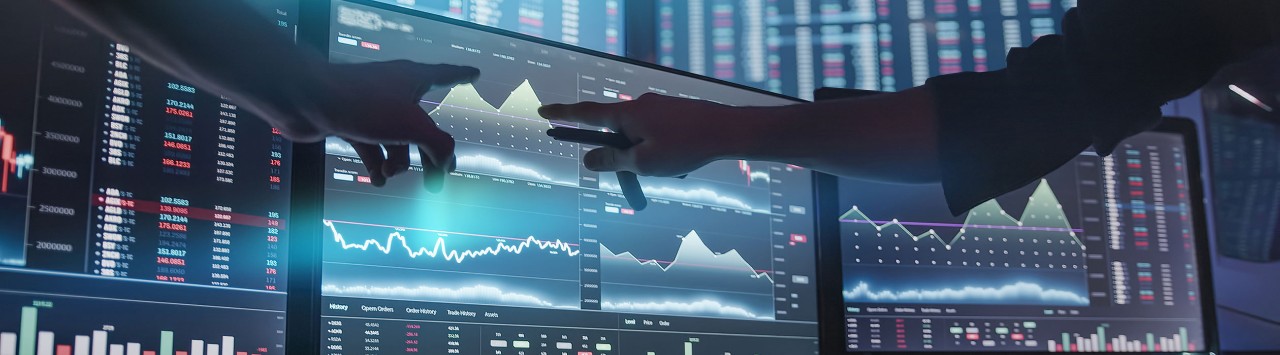 Two analysts pointing to digital monitors displaying financial data