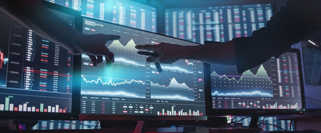 Two analysts pointing to digital monitors displaying financial data