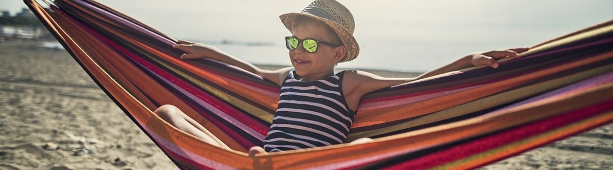 Young child wearing hat and sunglasses sitting on hammock at the beach
