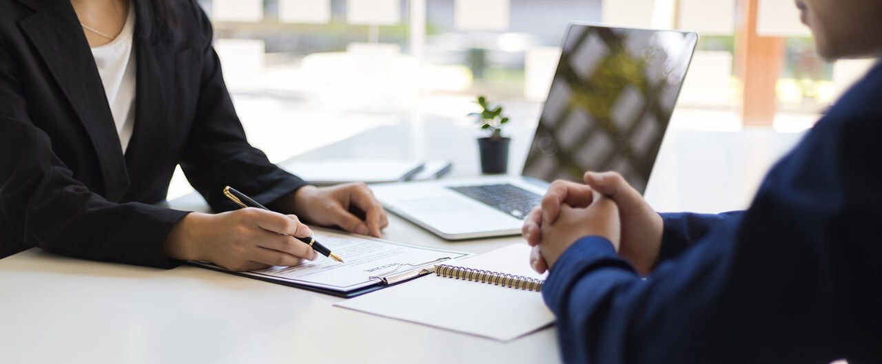 A man and a woman collaborate at a table with a laptop between them, discussing something on the screen.