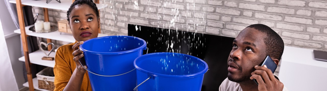 Couple using buckets to collect water from leaky ceiling while calling plumber