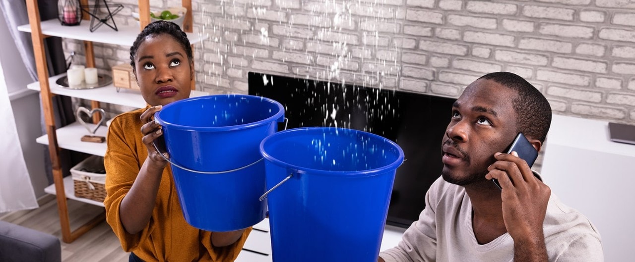 Couple using buckets to collect water from leaky ceiling while calling plumber