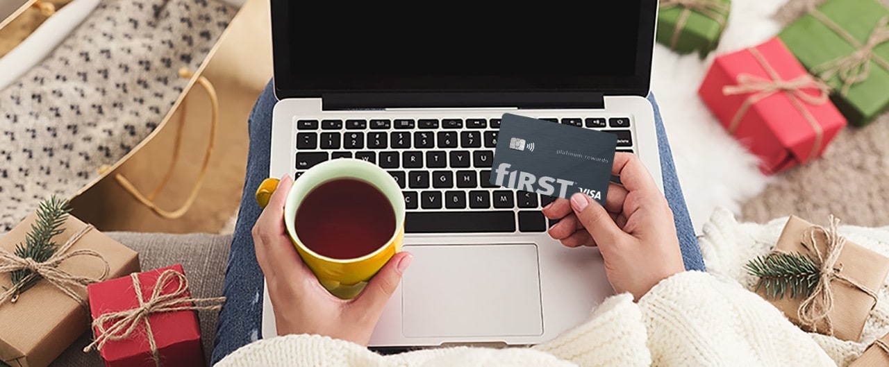 Woman holding First Financial Rewards card and a cup of coffee looking at laptop surrounded by holiday gifts