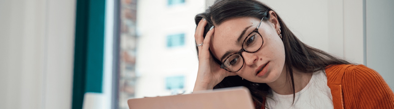 A concerned young woman wearing glasses looks at a laptop while resting her head in her hand.