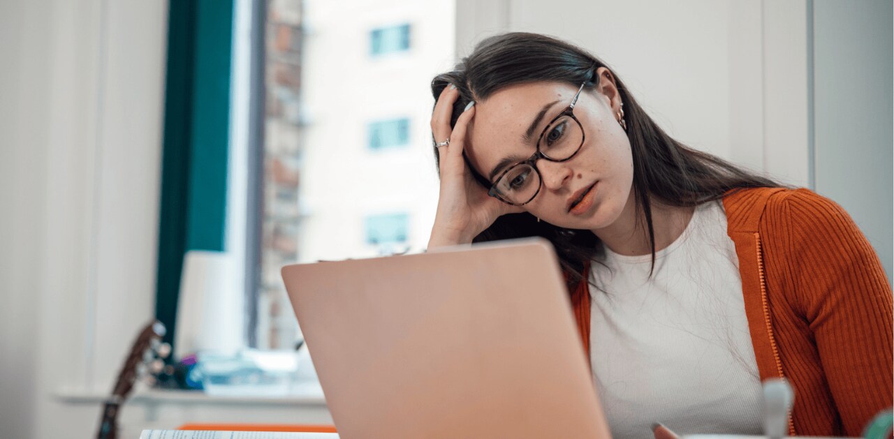 A concerned young woman wearing glasses looks at a laptop while resting her head in her hand.