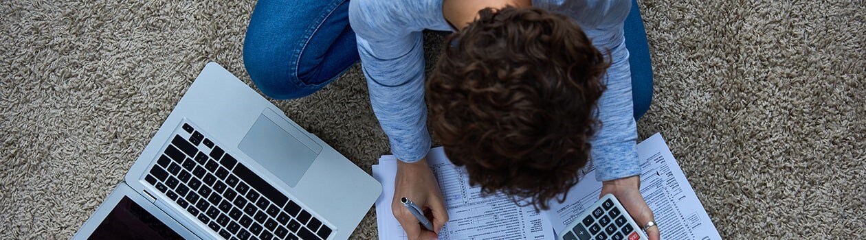 Overhead view of woman holding calculator, sitting on floor next to laptop preparing tax documents