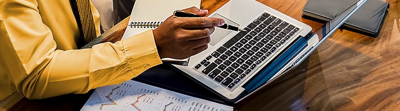 Man at desk with financial papers pointing pen at laptop