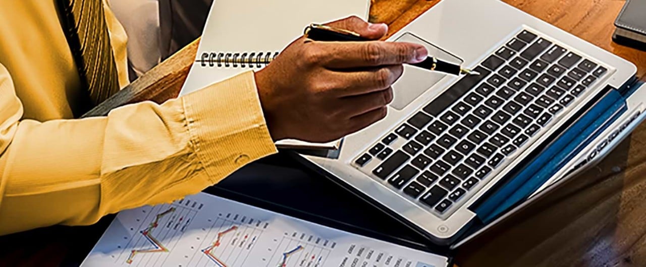 Man at desk with financial papers pointing pen at laptop