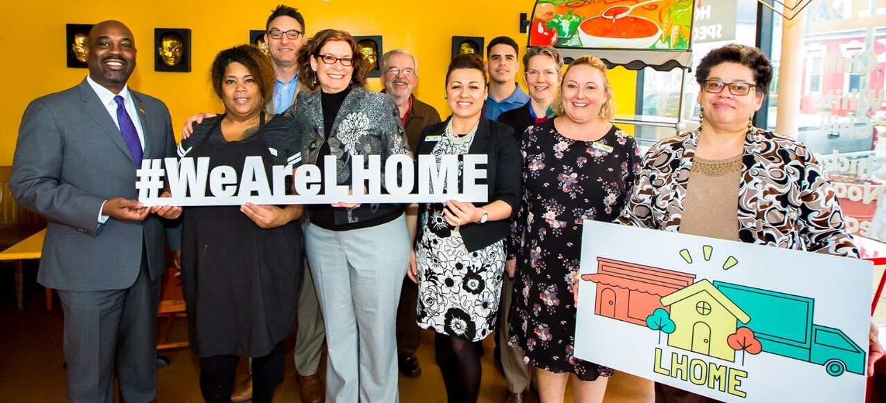 Diverse group of people holding a #WeAreLHOME sign and an LHOME sign