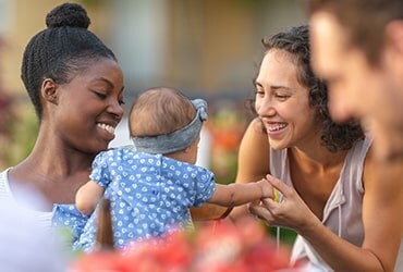 Black woman holding baby and a Caucasian woman holding baby's hand and playing with her