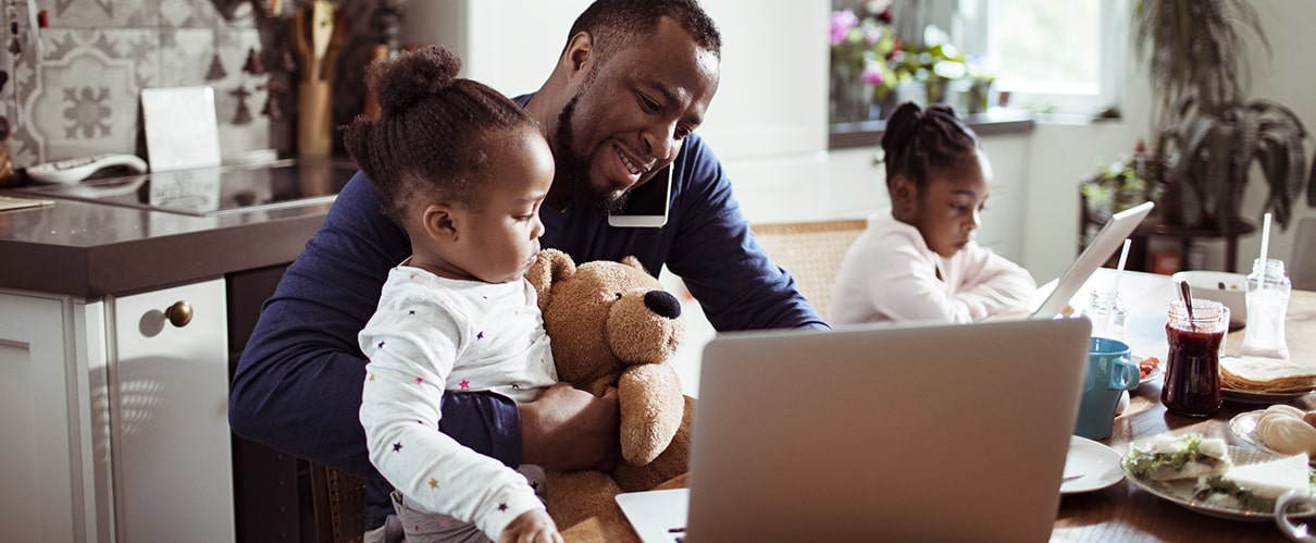 African-American father in front of laptop, talking on phone while holding young daughter, with older daughter sitting next to him